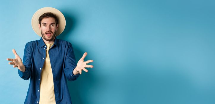 Surprised young man in summer hat, raising hands up staring with disbelief and amazement at camera, impressed with big event, standing on blue background.