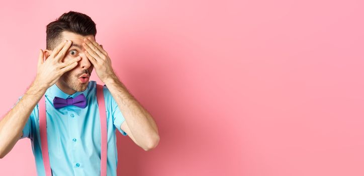 Intrigued funny guy in bow-tie, peeking through fingers at something interesting, checking out something cool, standing on pink background.