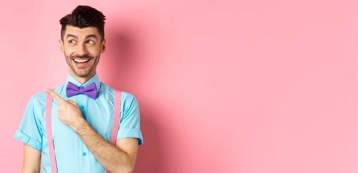 Cheerful caucasian man in funny bowtie looking and pointing hand left, showing promo logo, standing on pink background. Copy space