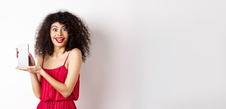 Fashionable lady in red dress and makeup, showing mobile phone screen and smiling, introduce smartphone application, standing over white background.