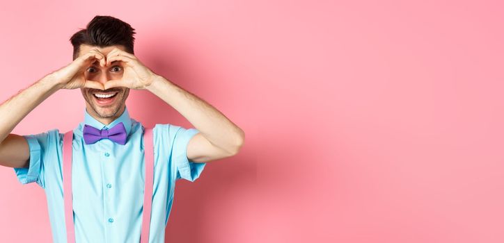 Happy Valentines day. Young man making hand heart and looking through it at camera, feeling romantic with lover, standing over pink background.