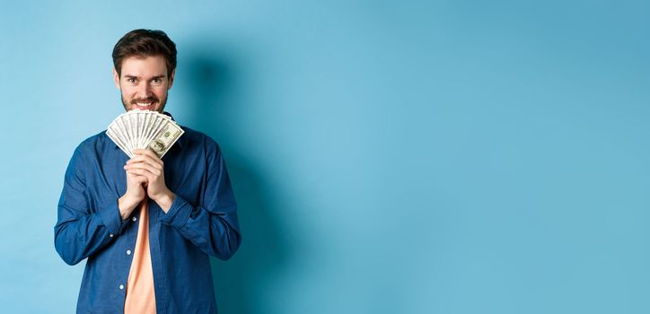 Happy smiling man holding money and looking at camera pensive, thinking of shopping, standing on blue background.