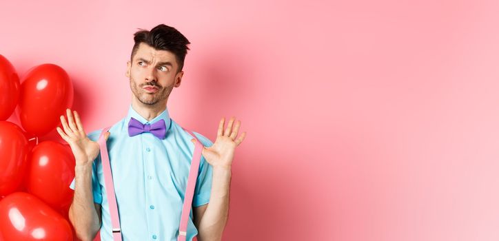 Valentines day concept. Pensive young man in bow-tie, raising hands up and looking left while thinking, making decision, standing on romantic pink background and heart balloons.