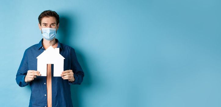 Real estate and quarantine concept. Happy young man in medical mask showing house cutout, buying property, standing on blue background.