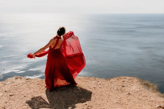 Side view a Young beautiful sensual woman in a red long dress posing on a rock high above the sea during sunrise. Girl on the nature on blue sky background. Fashion photo.