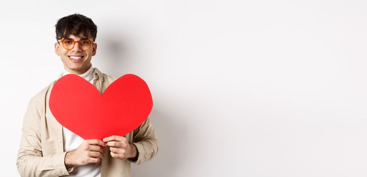 Handsome hipster guy with sunglasses and earring, waiting for true love on Valentines day, holding big red heart and smiling, standing over white background.