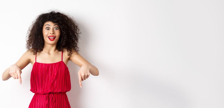 Excited woman with curly hair and red lips, wearing evening dress, gasping and pointing fingers down at super cool promo, standing on white background.