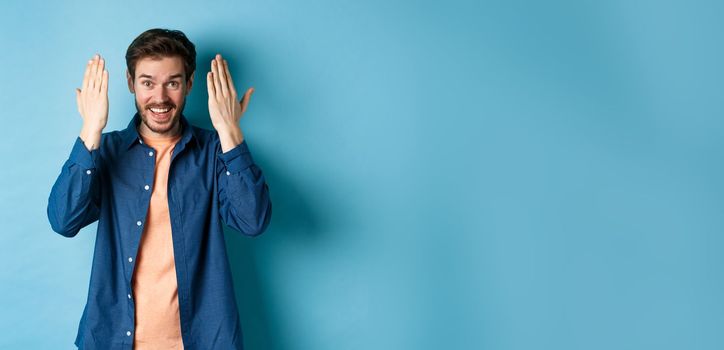 Happy young man open eyes and look surprised at gift, standing on blue background.