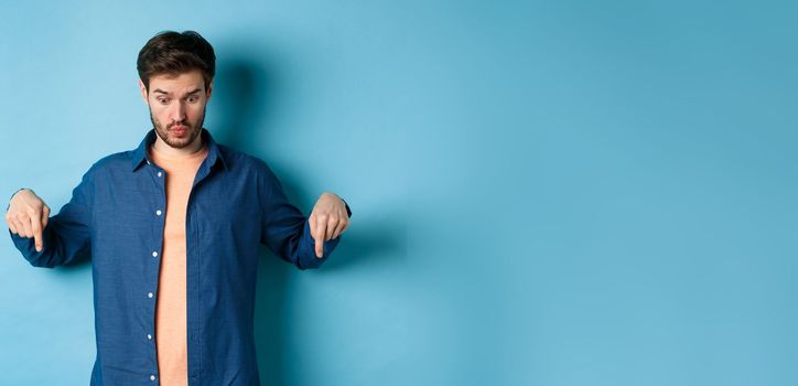 Image of shocked young man in casual clothes, looking and pointing down at something strange, standing on blue background.
