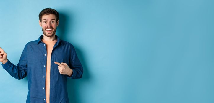 Excited and happy caucasian man smiling at camera, pointing fingers left at empty space, showing logo, standing on blue background. Copy space