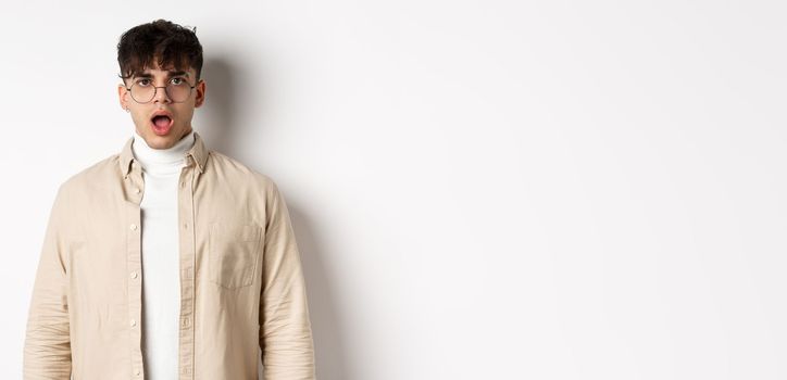 Portrait of shocked young man in glasses drop jaw, open mouth and stare amazed at camera, looking at something impressive, standing on white background.