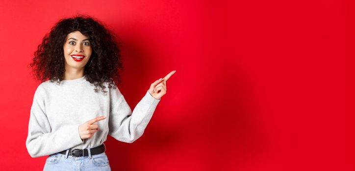 Amused young woman with curly hairstyle, pointing fingers right at logo, looking impressed and excited, checking out promotion, red background.