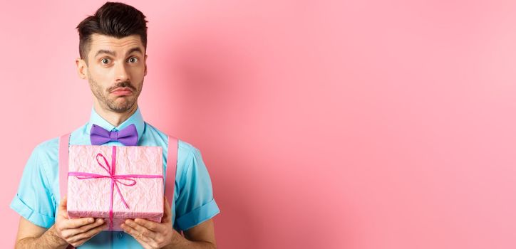 Holidays and celebration concept. Attractive young man with moustache, wearing festive outfit with bow-tie, showing cute gift box and looking at camera, standing over pink background.