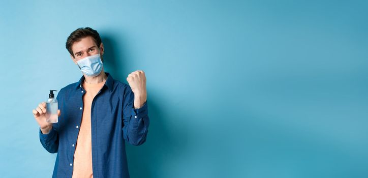 Coronavirus, quarantine and social distancing concept. Cheerful guy say yes and raise fist up while showing hand sanitizer, recommending product, blue background.