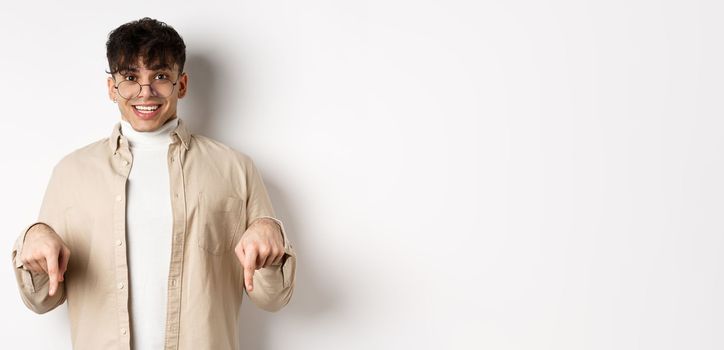 Portrait of happy young guy in glasses showing cool promo, pointing fingers down and smiling excited, standing on white background.