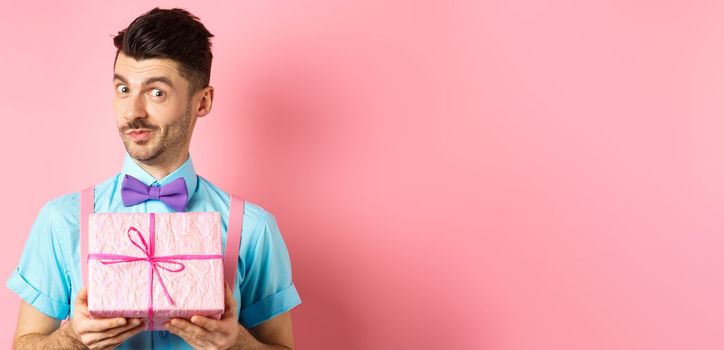 Holidays and celebration concept. Funny guy with moustache showing cute gift box wrapped in festive paper, looking at camera, presenting you, standing on pink background.