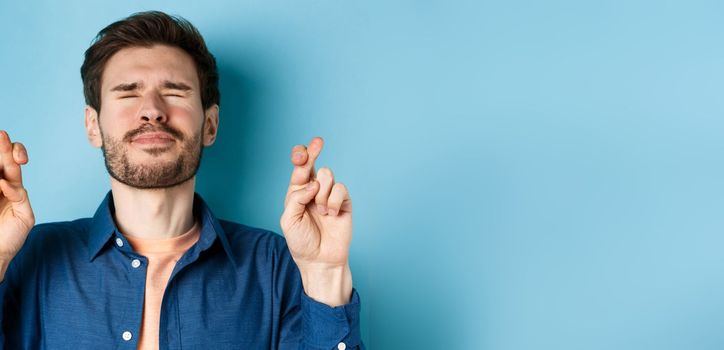 Close-up of modern young man making wish, close eyes and cross fingers for good luck, praying for dream come true, standing on blue background.