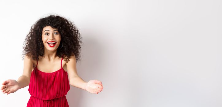 Portrait of happy caucasian woman in red dress and makeup, stretch out hands to beckon someone, inviting for hug, receiving surprise gift, standing on white background.