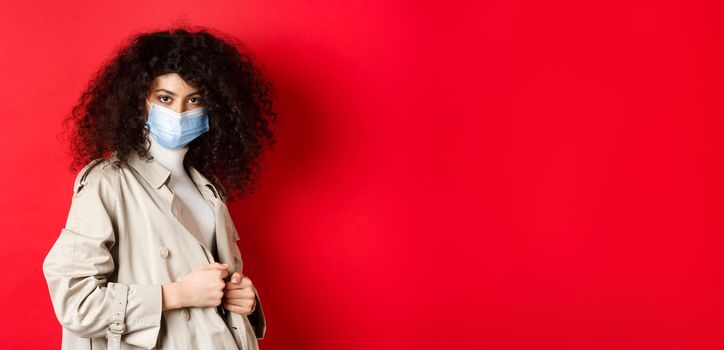 Covid-19, pandemic and quarantine concept. Sassy girl in trench coat and medical mask, put on trench coat for spring walk, red background.