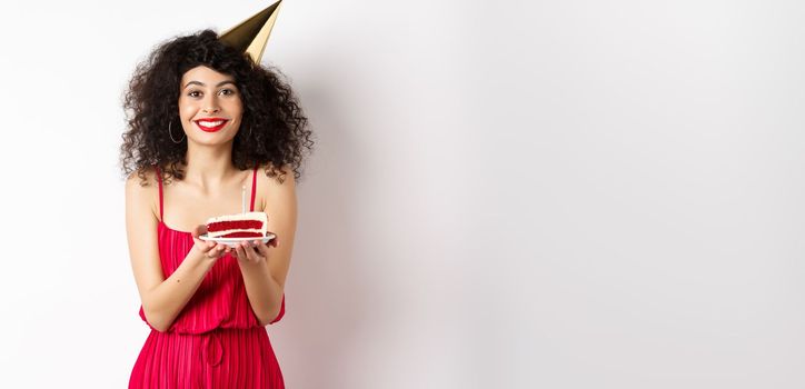 Beautiful woman in red dress, wearing party hat and celebrating birthday, holding b-day cake and making wish, smiling at camera, standing on white background.