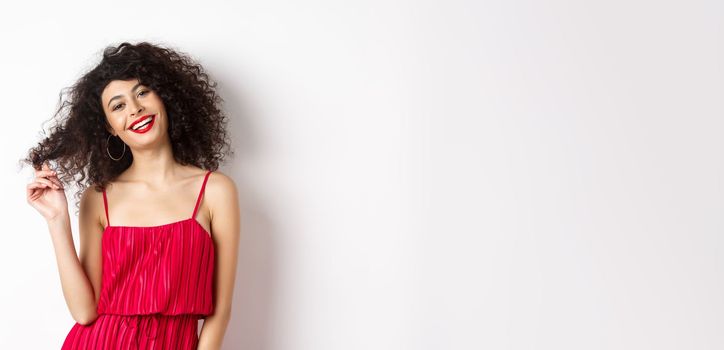 Beautiful woman with curly hair, wearing red dress and lipstick, playing with curl strand and smiling at camera, white background.