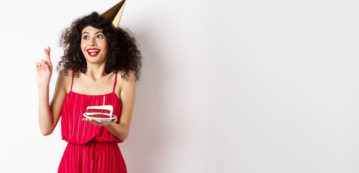 Excited birthday girl making wish, cross fingers good luck and looking aside at logo, celebrating bday, holding piece of cake, white background.