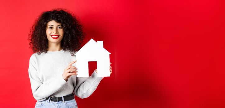 Real estate. Smiling caucasian woman with curly hair and red lips, showing paper house model, searching property, standing on red background.