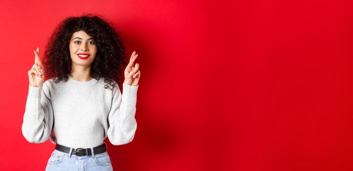 Hopeful young woman with red lips and curly hair, cross fingers for good luck and making wish, praying for dream come true, smiling excited, red background.