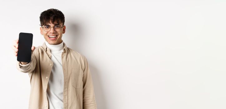 Handsome young man showing empty smartphone screen, standing on white background. Copy space