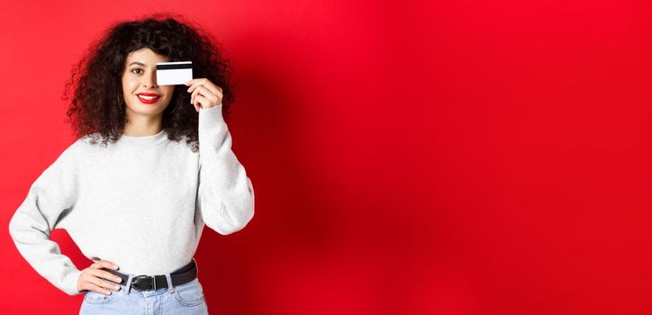 Beautiful female model with curly hair, showing plastic credit card and smiling, standing on red background. Makeup and shopping concept.