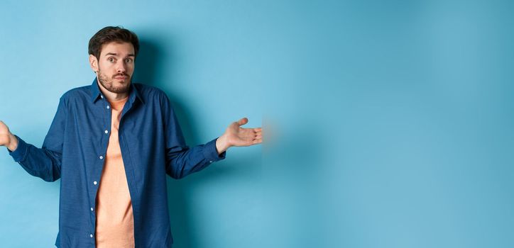 Confused young man with beard, shrugging shoulders and looking clueless at camera, know nothing, standing on blue background.