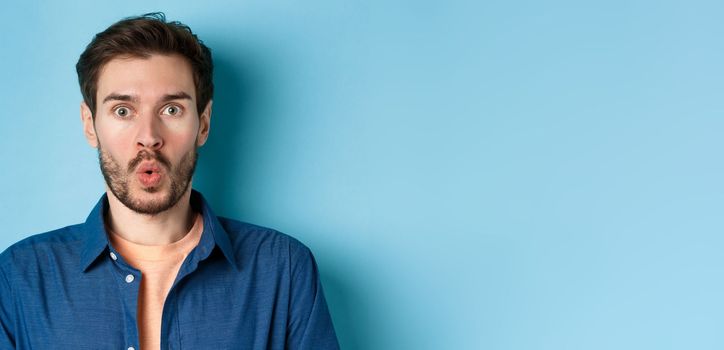 Close up portrait of amazed and surprised man saying wow, staring impressed at camera, checking out cool offer, standing on blue background.