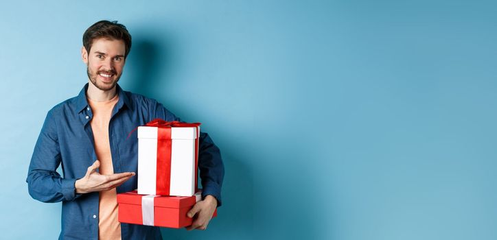 Young man in casual clothes buying romantic gifts on Valentines day, pointing at presents boxes and smiling, standing over blue background.