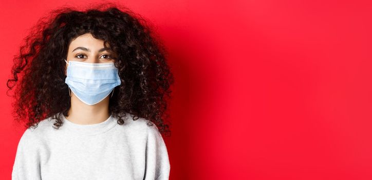 Covid-19 and pandemic concept. Close-up of modern young woman with curly hair, wearing medical mask from coronavirus, smiling at camera, red background.