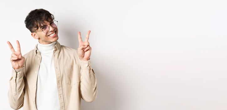 Real people. Handsome hipster guy in glasses showing peace gestures and smiling cute, standing on white background.