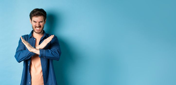 Angry young man frowning and clenching teeth outraged, showing cross gesture to stop or forbid something, standing on blue background.