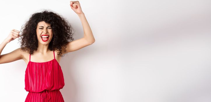 Cheerful emotive woman with curly hair, red dress, raising hands up and chanting, rooting for team, shouting wanting to win, standing on white background.