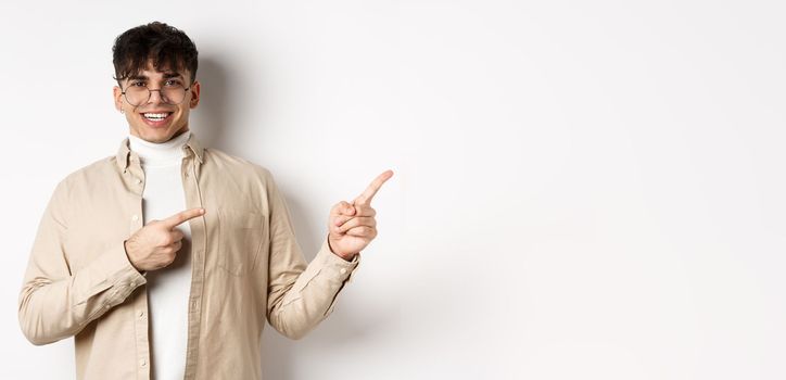 Proud and happy young man in glasses showing logo, pointing fingers at upper right corner and smiling, standing on white background.