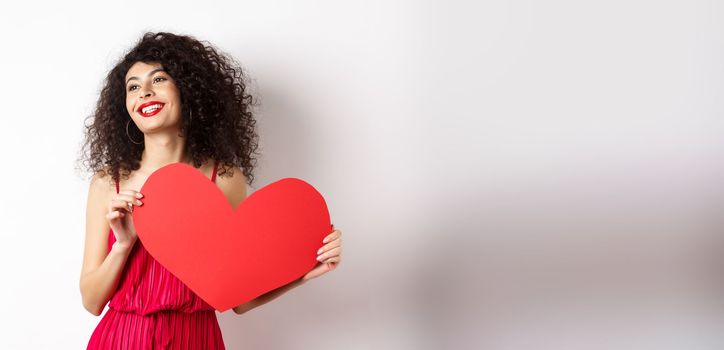 Romantic woman in dress and makeup, looking aside with dreamy face and love, showing big red valentines heart card, standing on white background.