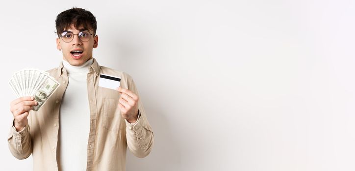 Excited young guy showing dollar bills and credit card, earn money and looking amazed, standing on white background.
