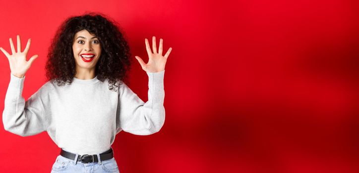 Happy beautiful woman smiling and raising hands up, showing number ten, ordern dozen of something, standing in sweatshirt on red background.