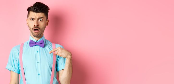 Image of confused funny guy in bow-tie and suspenders, pointing at himself as if being accused or chosen, raising eyebrow surprised, standing over pink background.