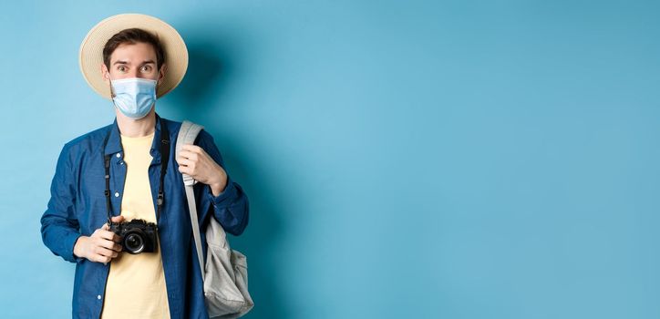 Covid-19 and travelling concept. Young guy tourist in medical mask and summer hat travel abroad during coronavirus pandemic, taking pictures on vacation, blue background.