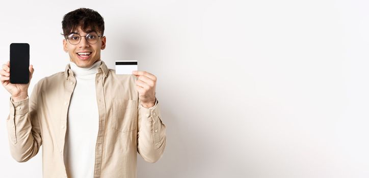 Online shopping. Surprised and happy young man showing credit card and mobile phone screen, standing on white background.