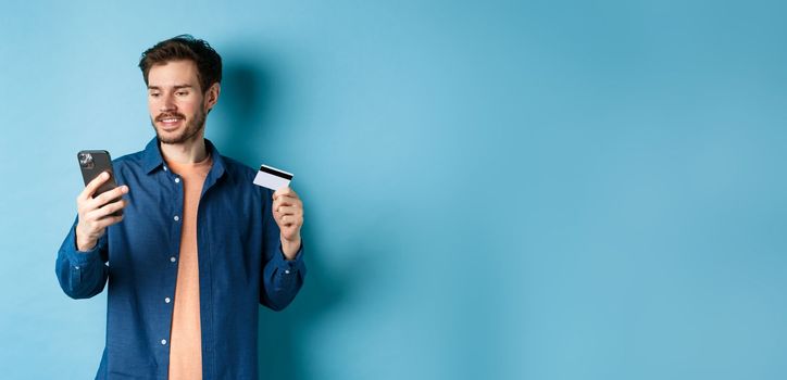 E-commerce concept. Image of handsome caucasian man paying online, looking at mobile phone screen and holding plastic credit card, blue background.