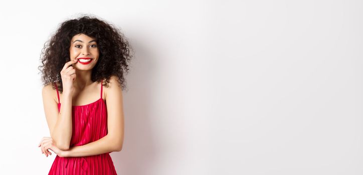 Excited beautiful woman in red dress, biting finger and smiling cute at camera, want something, standing on white background.