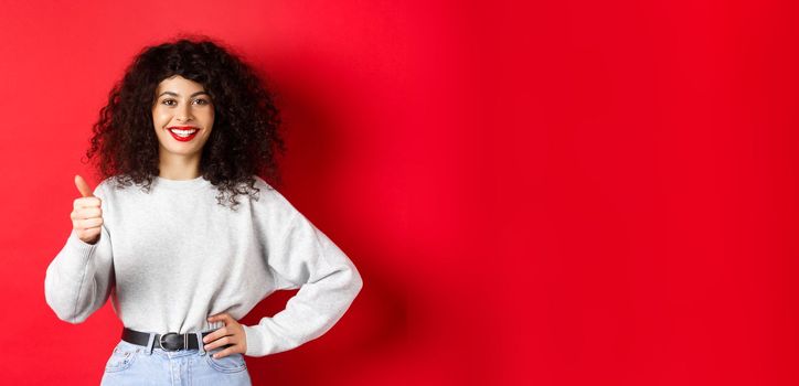 Enthusiastic young woman with curly hair, red lips, showing thumbs up and smiling in approval, praise good product, standing against red background.