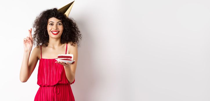 Excited birthday girl celebrating, making wish, holding cake and cross fingers good luck, standing happy on white background.