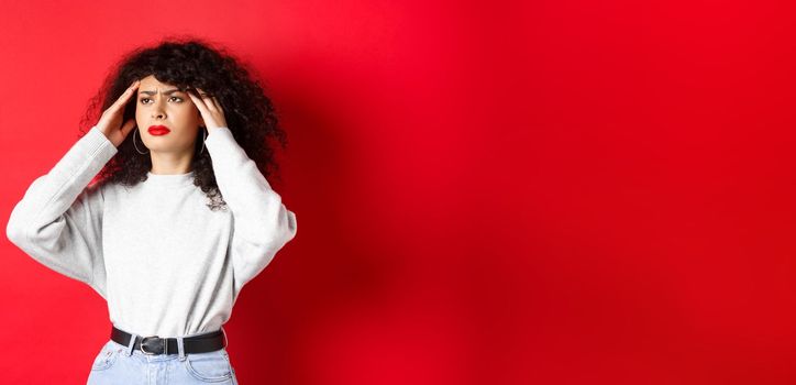 Distressed italian girl with curly hair, touching head temples and feeling bad, having headache, looking aside, standing on red background.