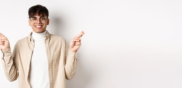 Handsome and positive young man smiling, making wish with crossed fingers and happy face, hope for dream to come true, standing on white background.